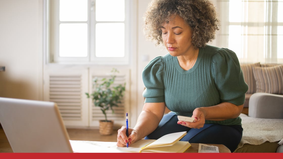 woman sitting on a sofa writing in a notebook with an open laptop on the coffee table